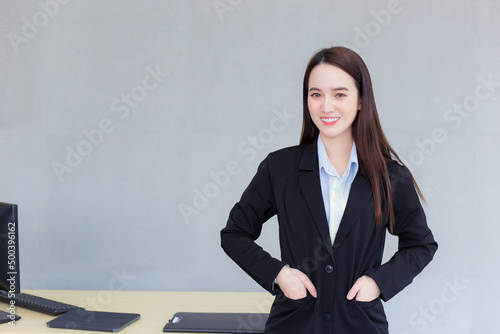 Professional business working Asian woman stands and put her hand in pocket of a black suit while she smiles happily in office as background.