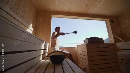 Handsome young man pouring water onto hot stone in the sauna
