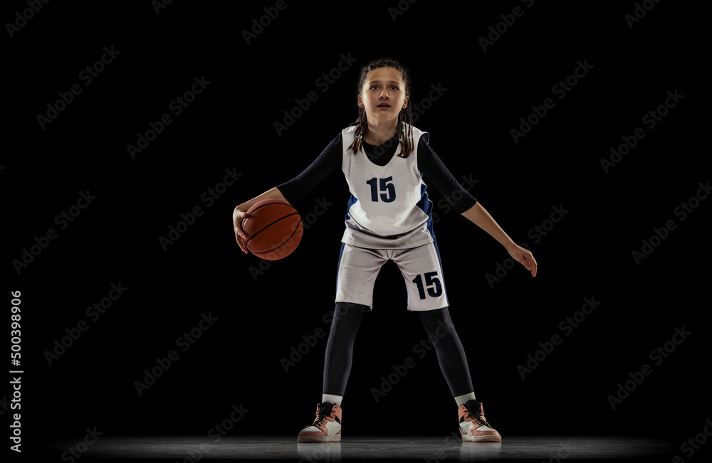 Portrait of teen girl, basketball player in motion, dribbling ball isolated over black studio background. Beat the defender