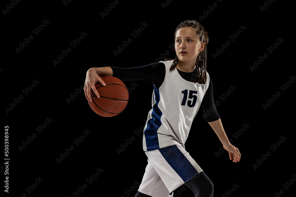 Training. Studio shot of teen girl in white uniform playing basketball, doing dribbling exercise isolated over black background.