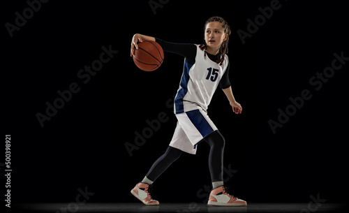 Portrait of teen girl, basketball player in motion, dribbling ball isolated over black studio background