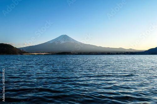 A close up view on Mt Fuji from the side of Kawaguchiko Lake, Japan. Soft colors of sunset - golden hour. Top of the volcano covered with a snow layer. Serenity and calmness. Calm lake's surface © Chris