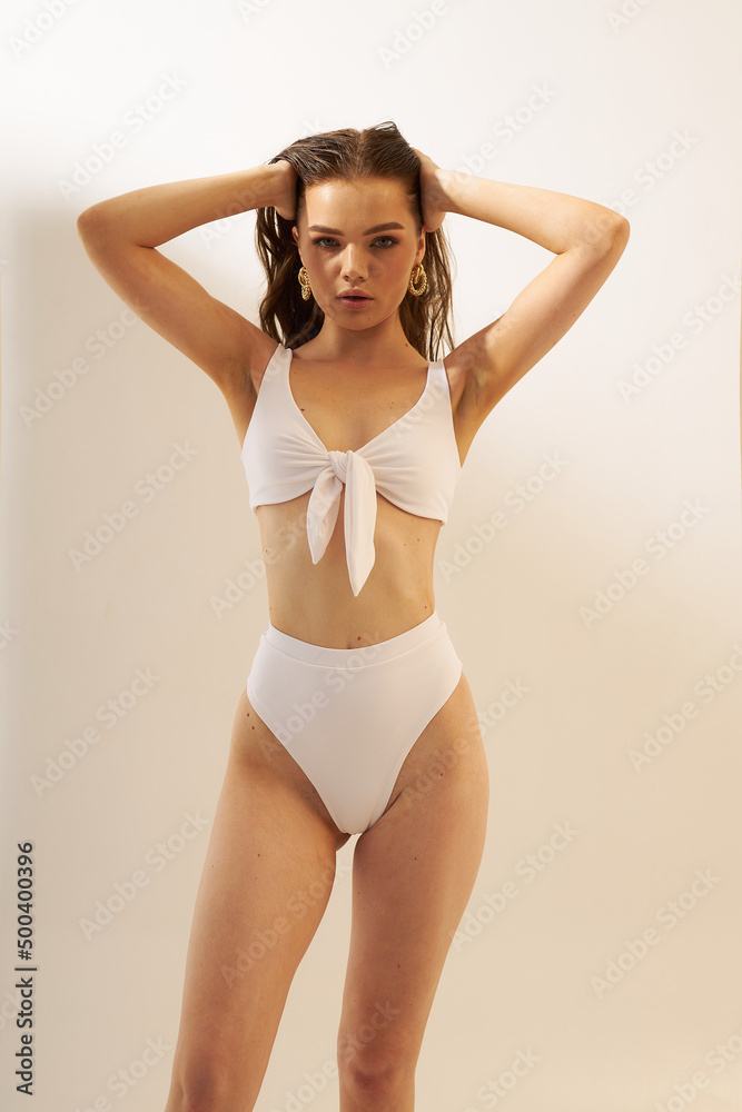 Obraz premium Fashion photo of young lady with perfect body in a white designer trend 2022 swimsuit. Girl on a sunny day sunbathes in sexy pose on the white. Sun screen, summer concept.
