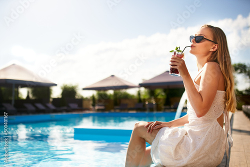 Blonde woman drinks refreshing cocktail sunbathing and sitting near swimming pool at tropical spa. Female in sunglasses enjoys a drink on poolside. Girl chilling in tropical resort on vacation.