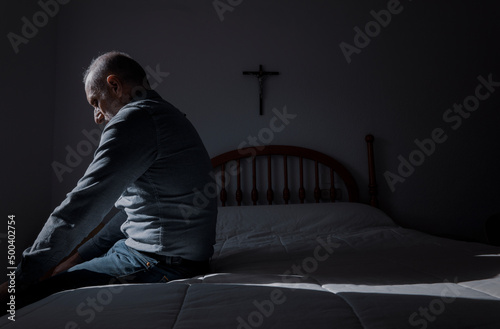 Side view of adult man sitting on bed with religious cross on wall © WeeKwong