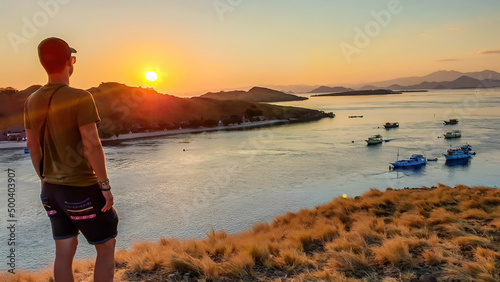 A man standing on top of a small island, facing a rising sun over Komodo National Park, Flores, Indonesia. Golden hour over the islands and sea. Some boats anchored to the bay. New day begining photo