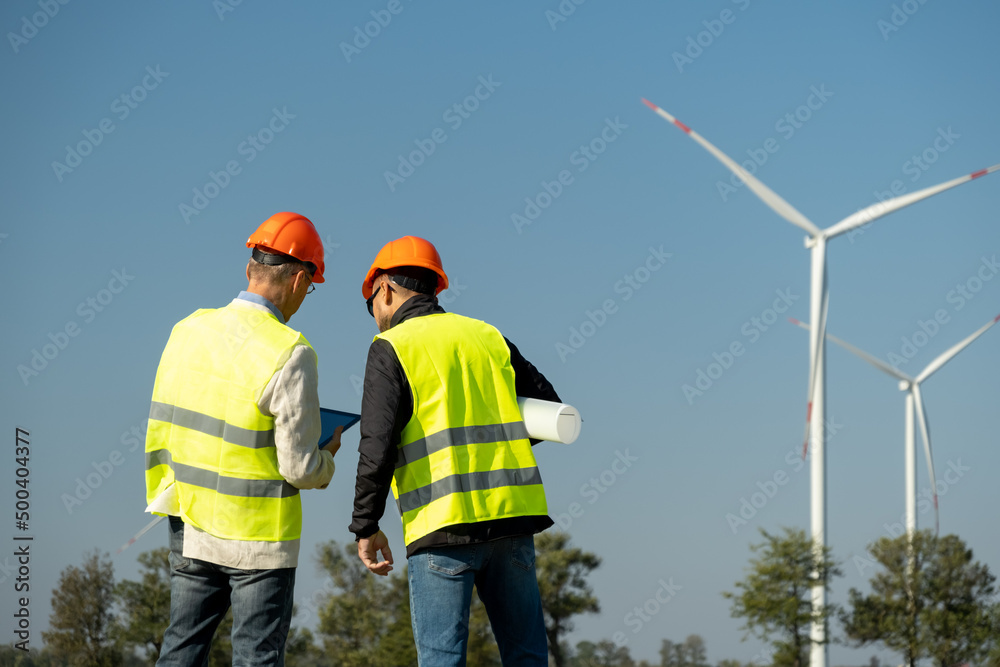 Wind turbines generate electric energy in front of engineers. Engineers in protective helmets stand holding project papers roll against rotating windmills