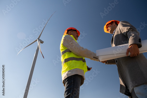 Wind turbines generate alternative electric energy. Supervisor with project papers shakes hands with general engineer side low angle shot