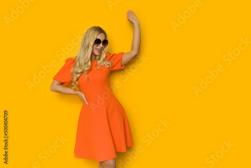 Happy blond woman in orange cocktail dress is looking to the side and waving hand.
