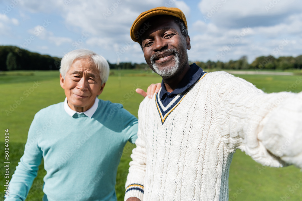 Φωτογραφία smiling african american man taking selfie with senior asian friend of golf field