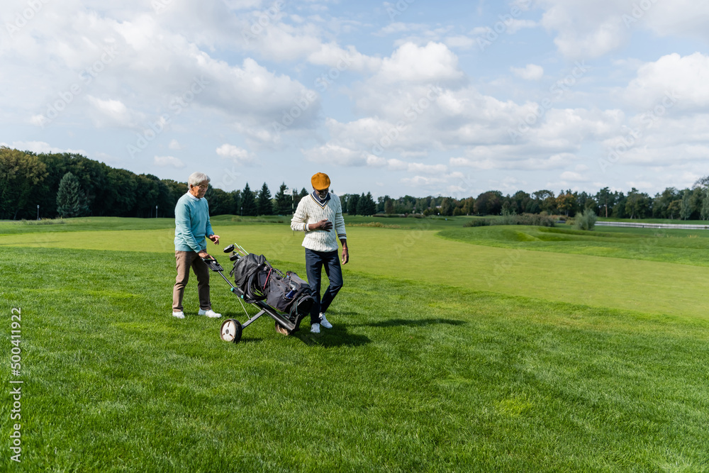 senior asian man walking with golf cart near wealthy african american friend. senior asian man walking with golf cart near wealthy african american friend.