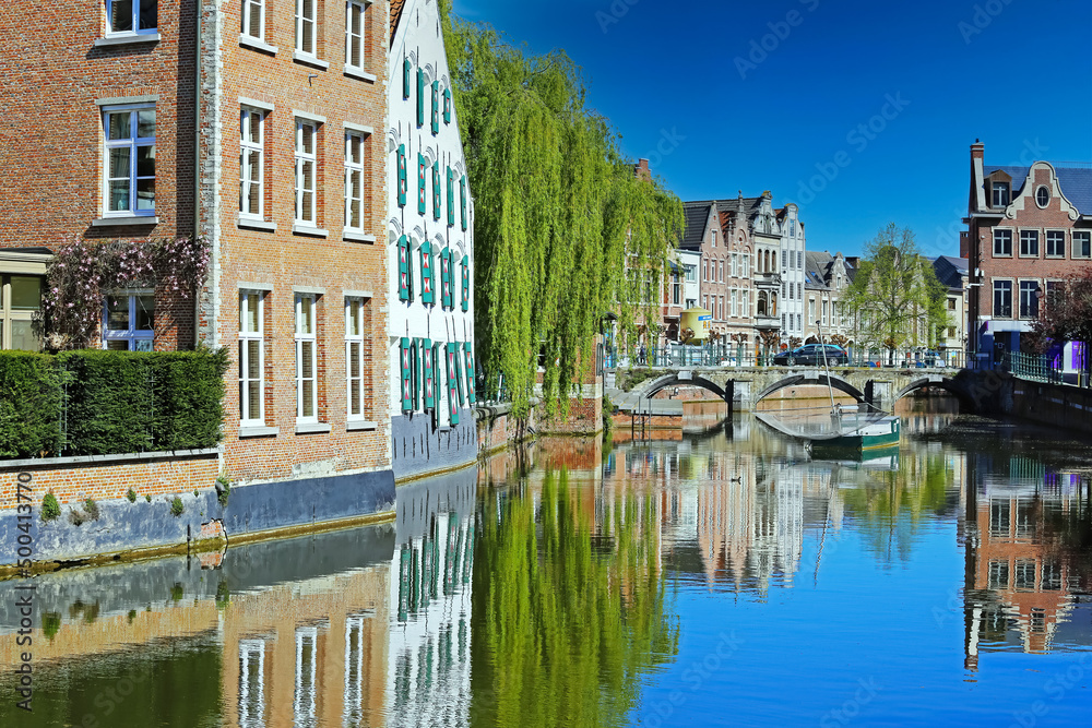 Lier, Belgium - April 9. 2022: View over water village moat on ancient houses, green weeping willow tree, medieval stone arch bridge, blue clear sky