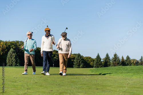 senior interracial friends walking with golf clubs on green field.