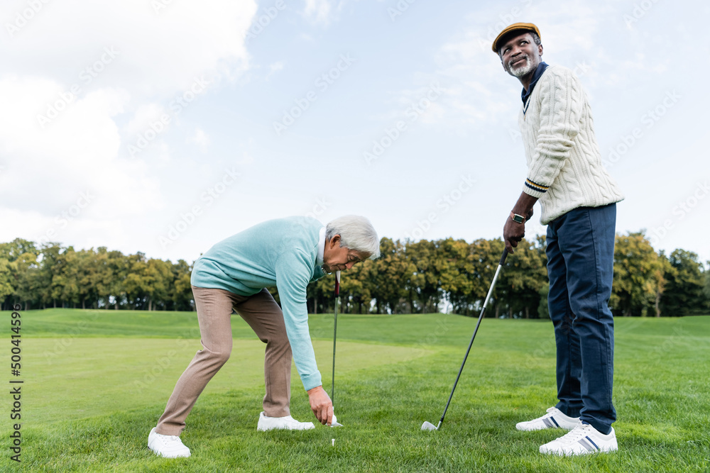 asian senior man putting ball on golf tee near african american friend. asian senior man putting ball on golf tee near african american friend.