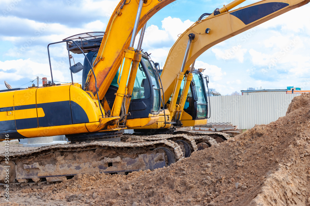Two powerful excavators work at the same time on a construction site, sunny blue sky in the background. Construction equipment for earthworks. Two powerful excavators work at the same time on a construction site, sunny blue sky in the background. Construction equipment for earthworks.