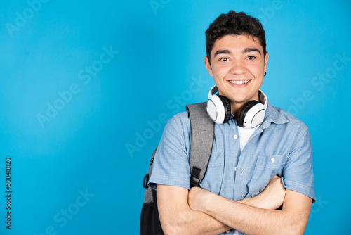 Hispanic college student looking at camera isolated on blue background. Portrait of teenager boy arms crossed