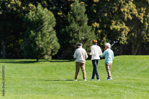 back view of senior multiethnic friends walking with golf clubs on green field near trees.