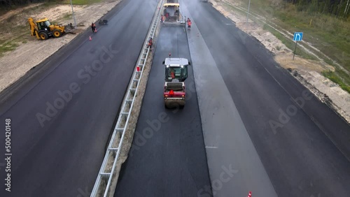 A road roller compacts the new blacktop on the motorway. Track is covered in steam from the hot asphalt. Work of heavy road machinery. Repair and renewal of the highway surface. Road workers on shift.