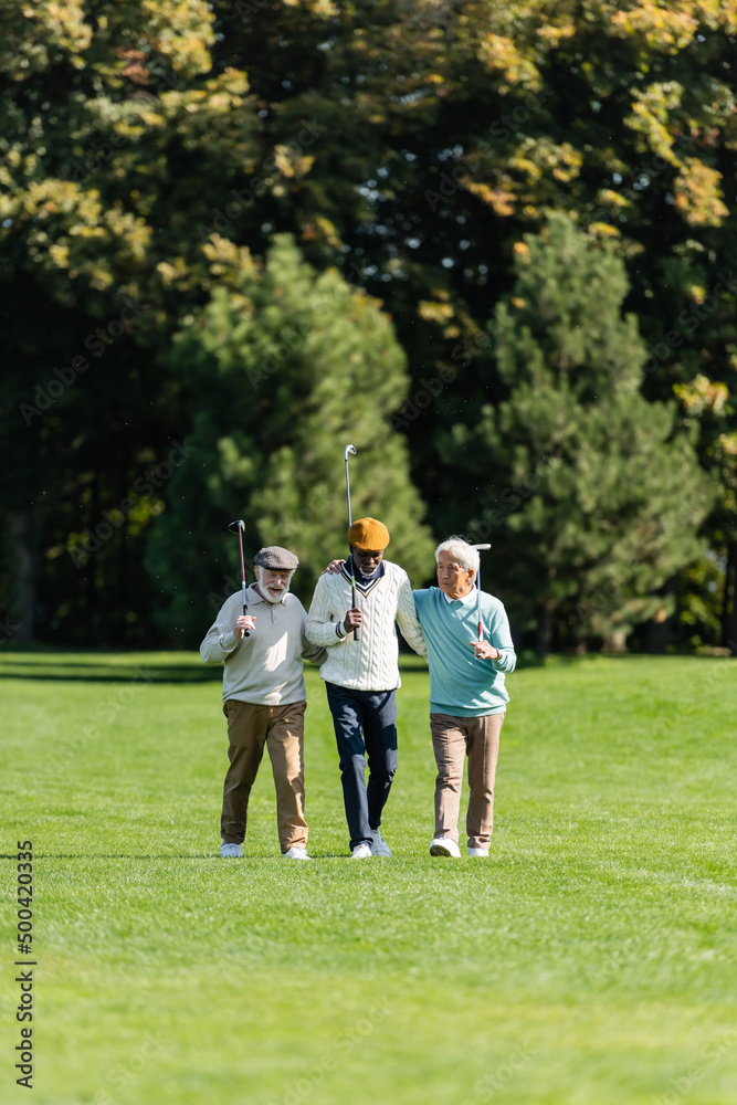 senior multiethnic friends walking with golf clubs on green field near trees.