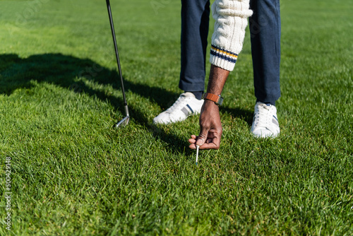 cropped view of african american senior man placing golf tee on green lawn.