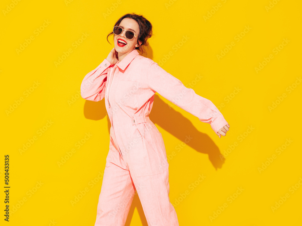Young beautiful smiling female in trendy summer pink overalls. Sexy carefree woman posing near yellow wall in studio. Positive model having fun and going crazy. Cheerful and happy. Dancing