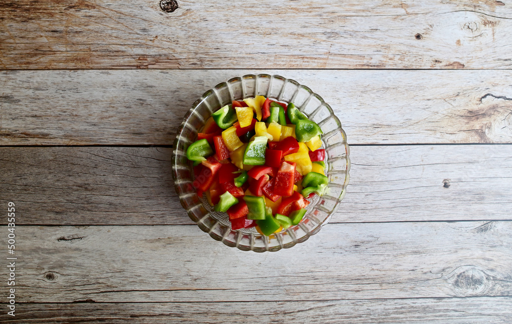 Top Views of Fresh red, green and yellow bell peppers (capsicum) are cut into small pieces. in a clear glass bowl laying on the wooden floor background. Concept for health and vitamins. Top Views of Fresh red, green and yellow bell peppers (capsicum) are cut into small pieces. in a clear glass bowl laying on the wooden floor background. Concept for health and vitamins.