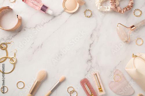 Top view photo of pink barrettes rose quartz roller gua sha glass bottle eyeshadow scrunchies gold earrings rings wristlet and cosmetic bag on white marble background with copyspace in the middle