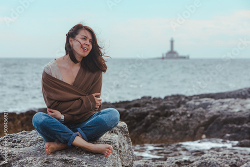 woman sitting by rocky sea beach in wet jeans lighthouse on background