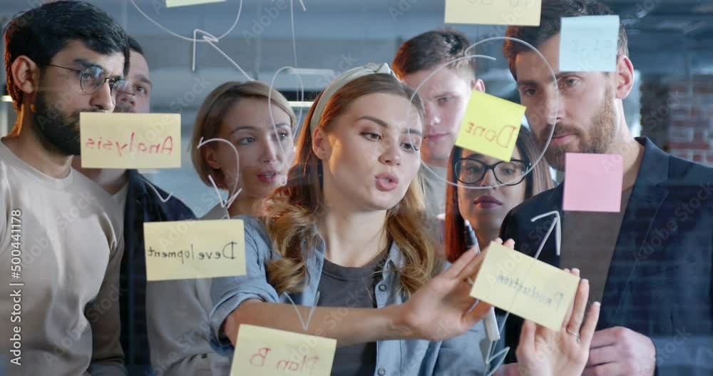 Young woman with her multiethnic working team making plans for company future and sticking to-do notes. Mixed-races males and females emplyees brainstorming and planning work for startup project.