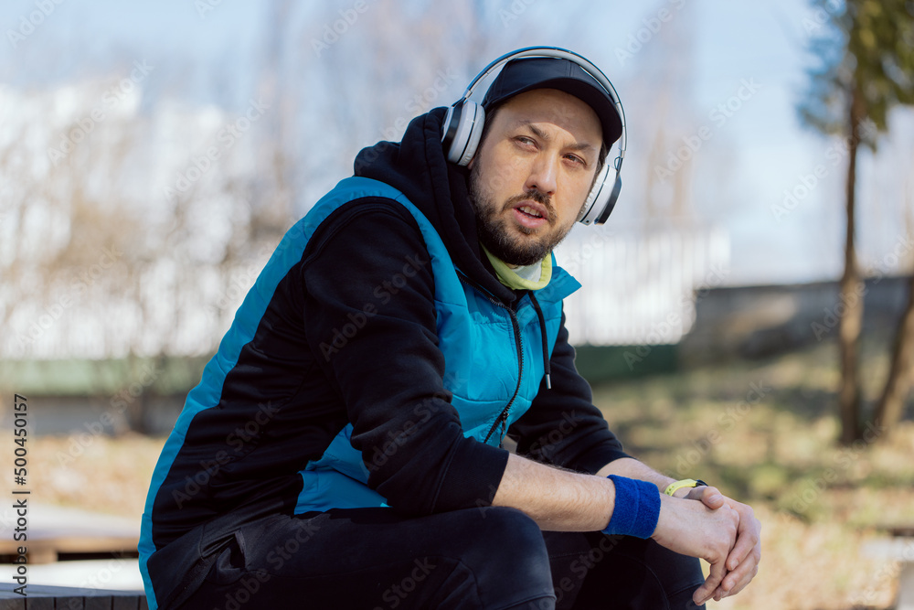 A thoughtful man sits on a wall in a park wearing sportswear and wireless headphones listening to music, turning on a workout on smartwatch, relaxing in the fresh air. A thoughtful man sits on a wall in a park wearing sportswear and wireless headphones listening to music, turning on a workout on smartwatch, relaxing in the fresh air.