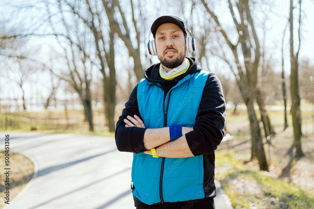Portrait of a smiling disciplined man in fitness attire running in a park on a spring morning. Sportsman in wireless headphones hands with wristbands crossed. Portrait of a smiling disciplined man in fitness attire running in a park on a spring morning. Sportsman in wireless headphones hands with wristbands crossed.