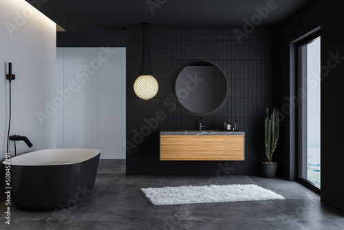 Modern bathroom interior with concrete floor, black bathtub, and oval mirror, marble basin, pendant light, cactus, front view. Minimalist black bathroom with modern furniture. 3d rendering 