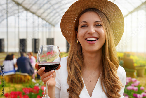 Close-up of young woman looking at camera and holding glass of wine with people among flowers field drinking and chatting on the background. Copy space.