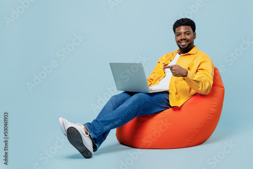 Full body happy young man of African American ethnicity wear yellow shirt sit wearing bag chair hold use work point finger on laptop pc computer isolated on plain pastel light blue background studio
