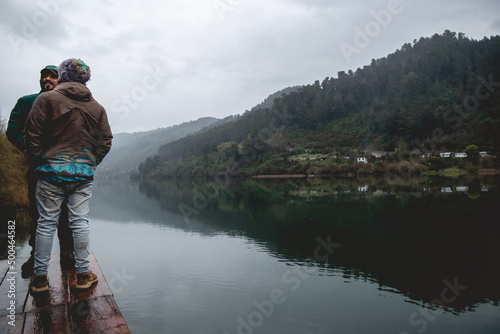 Two happy, handsome, young and bearded brother with wool cap and jackets smiling in a wood dock with view of lake in the mountains with green forest, mist, reflections in the water and cloudy sky