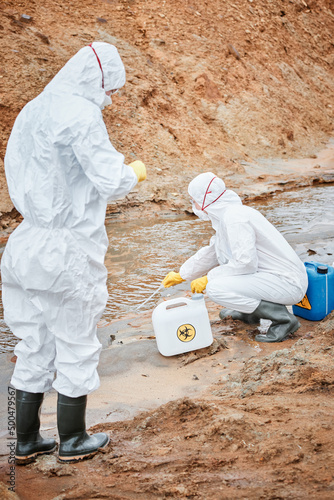 Ecological researchers in hazmat suits and rubber boots taking dirty water substance with pipette while studying it on polluted territory