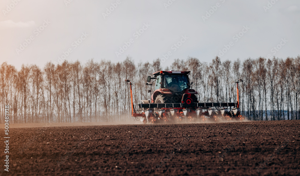 Fototapeta premium Spring sowing season. Farmer with a tractor sows corn seeds on his field. Planting corn with trailed planter. Farming seeding. The concept of agriculture and agricultural machinery.