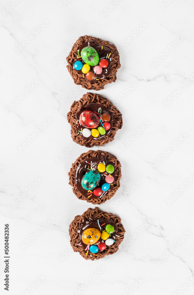 Easter chocolate cookie in the shape of a nest, decorated with a quail egg and colorful candy dragees and confectionery sprinkles. White background. Top view