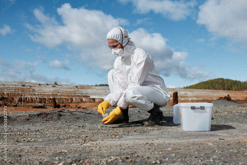 Ecologist in hazmat suit, mask, goggles and gloves gathering sample of soil on infertile land after environmental catastrophe Ecologist in hazmat suit, mask, goggles and gloves gathering sample of soil on infertile land after environmental catastrophe