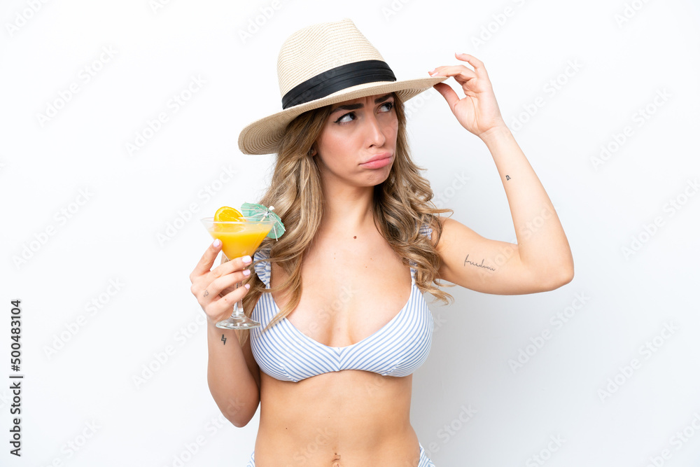 Young woman holding cocktail and wearing a bikini isolated on white background having doubts and with confuse face expression Young woman holding cocktail and wearing a bikini isolated on white background having doubts and with confuse face expression