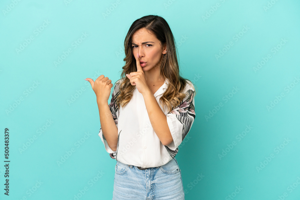 Young woman over isolated blue background pointing to the side and doing silence gesture Young woman over isolated blue background pointing to the side and doing silence gesture