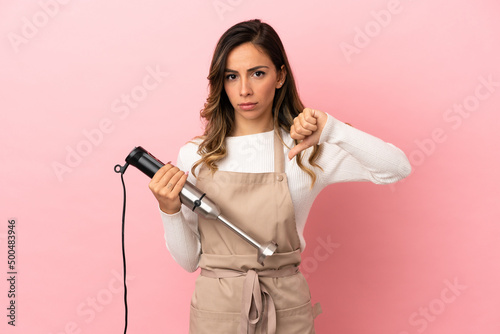Young woman using hand blender over isolated pink background showing thumb down with negative expression