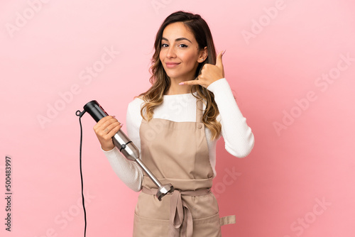 Young woman using hand blender over isolated pink background making phone gesture. Call me back sign