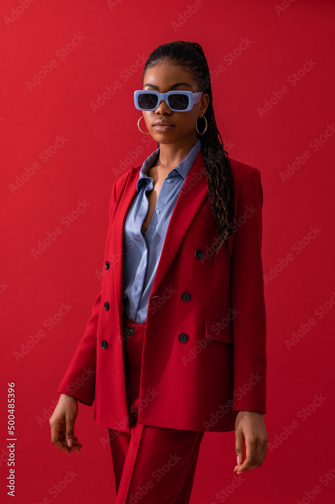 Fashionable Black woman wearing classic red suit with double breasted  blazer, satin shirt, trendy blue frame rectangle sunglasses. Fashion studio  portrait Stock Photo