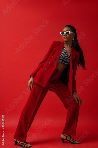 Elegant fashionable Black woman wearing classic red suit with blazer and trousers, zebra print top, trendy white frame sunglasses, strap sandals. Fashion full-length studio portrait 
