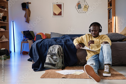 Serious African-American student guy in headphones sitting on floor in room with neon sticks and drawing illustration in sketchpad