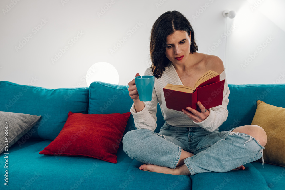 Woman reading a book and drinking tea while relaxing on a sofa at home Woman reading a book and drinking tea while relaxing on a sofa at home