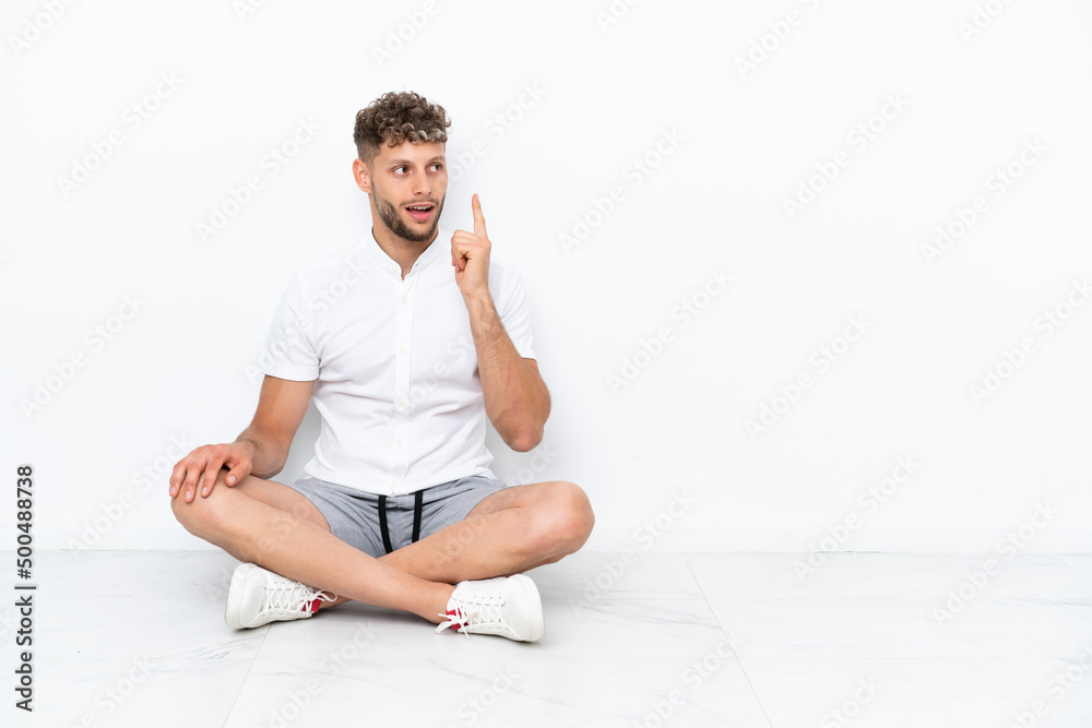 Fototapeta premium Young blonde man sitting on the floor isolated on white background thinking an idea pointing the finger up
