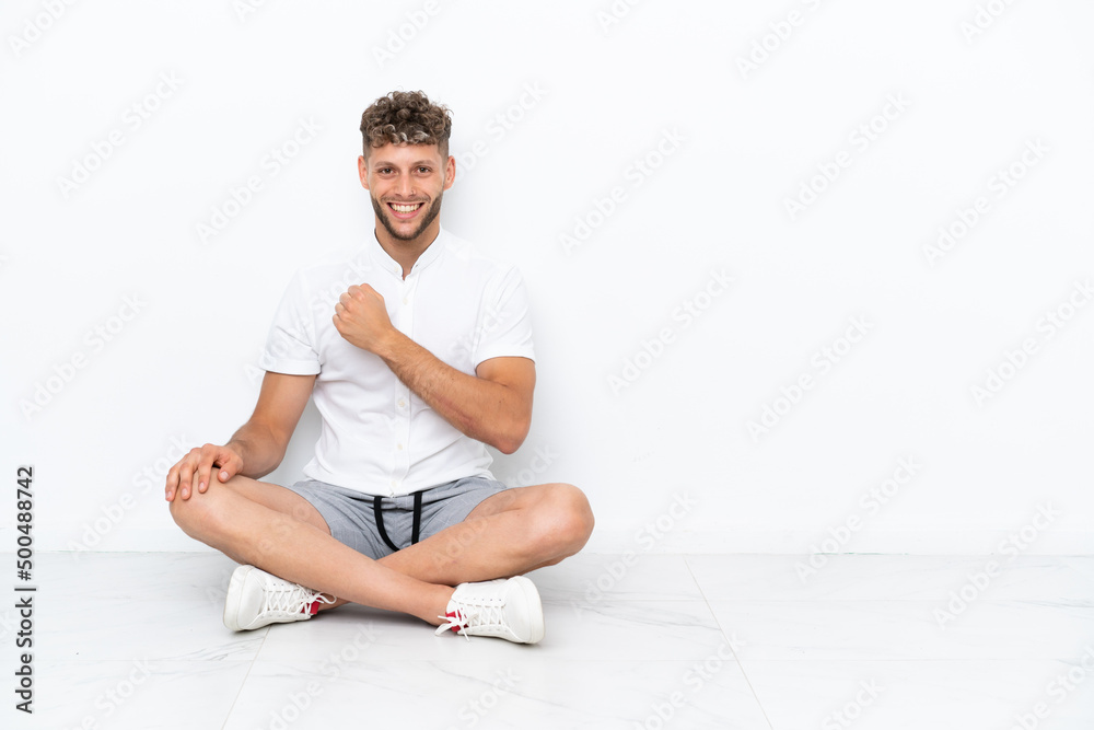 Young blonde man sitting on the floor isolated on white background celebrating a victory Young blonde man sitting on the floor isolated on white background celebrating a victory