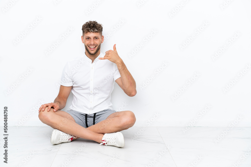Young blonde man sitting on the floor isolated on white background making phone gesture. Call me back sign Young blonde man sitting on the floor isolated on white background making phone gesture. Call me back sign